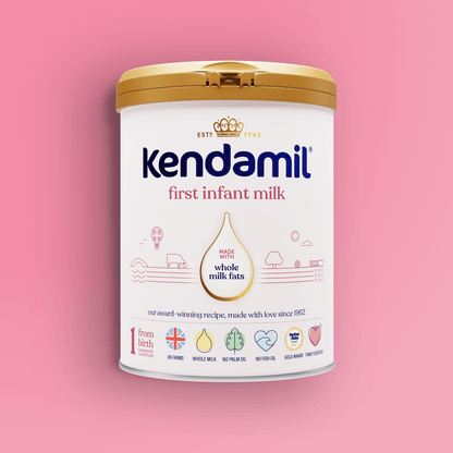 A tin of Kendamil Classic First Infant Milk with a gold lid sits on a pink background. The label highlights whole milk fats from grass-fed cows, UK origins, and icons for key ingredients and benefits for infants from birth.