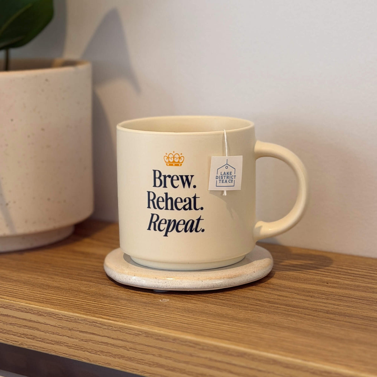 A “Brew, Reheat, Repeat.” Mug in a cream color rests on a coaster atop a wooden shelf. The durable mug features a tea bag label hanging over its edge, while a potted plant is partially visible in the background.