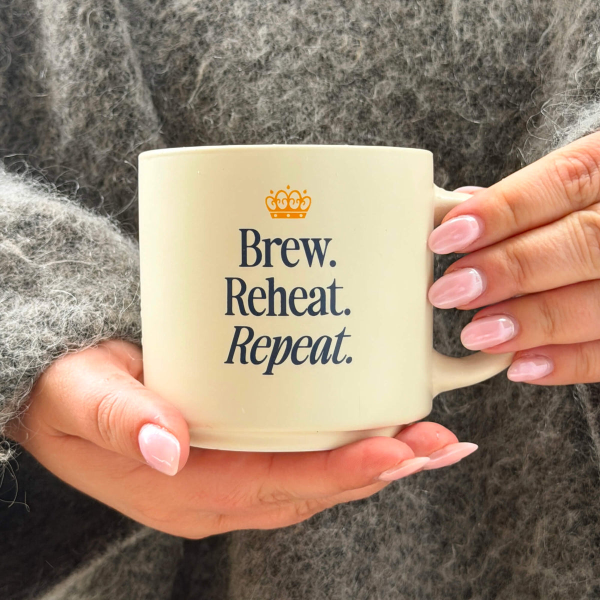 A person with light pink nails and a gray fuzzy sweater holds the “Brew, Reheat, Repeat.” Mug, which features a small crown icon above the text.