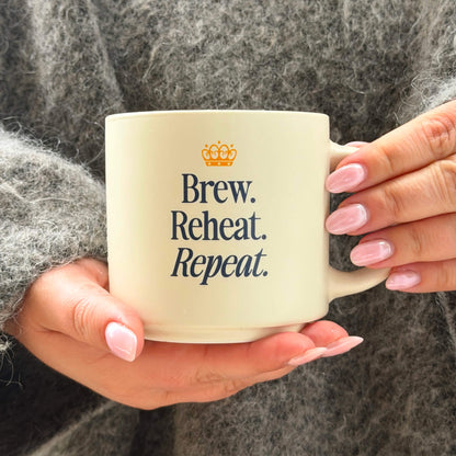 A person with light pink nails and a gray fuzzy sweater holds the “Brew, Reheat, Repeat.” Mug, which features a small crown icon above the text.