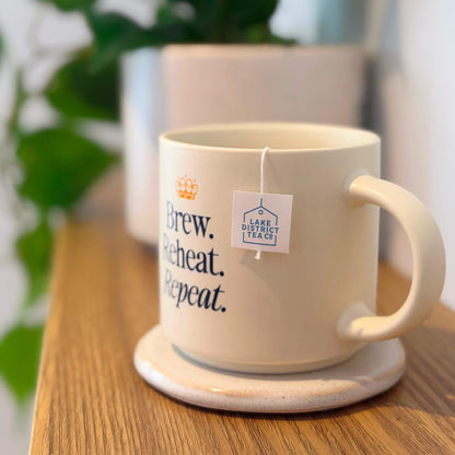 A sturdy “Brew, Reheat, Repeat.” Mug sits on a coaster beside a Lake District Tea Co. teabag, with a blurred plant and another pot visible in the background.