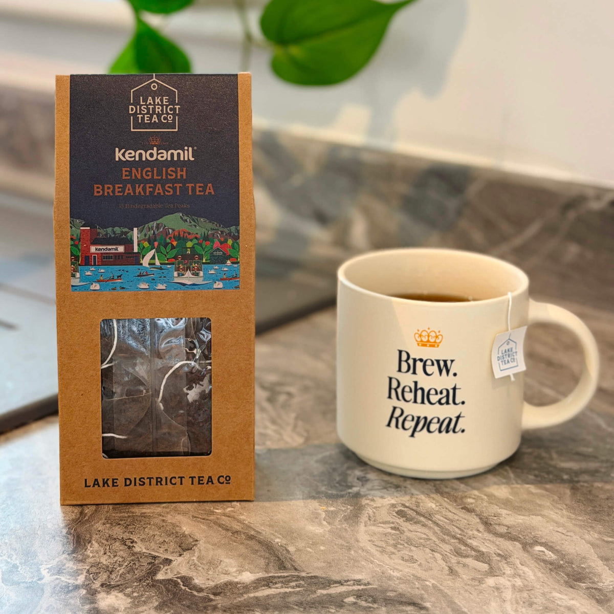 A box of Kendamil x Lake District Breakfast Tea stands beside a white mug featuring the words Brew. Reheat. Repeat. on a marble countertop, with a tea bag tag draped over the rim. In the background, a green plant accents this rich, full-bodied tea blended from Assam, Ceylon, and Kenyan leaves.