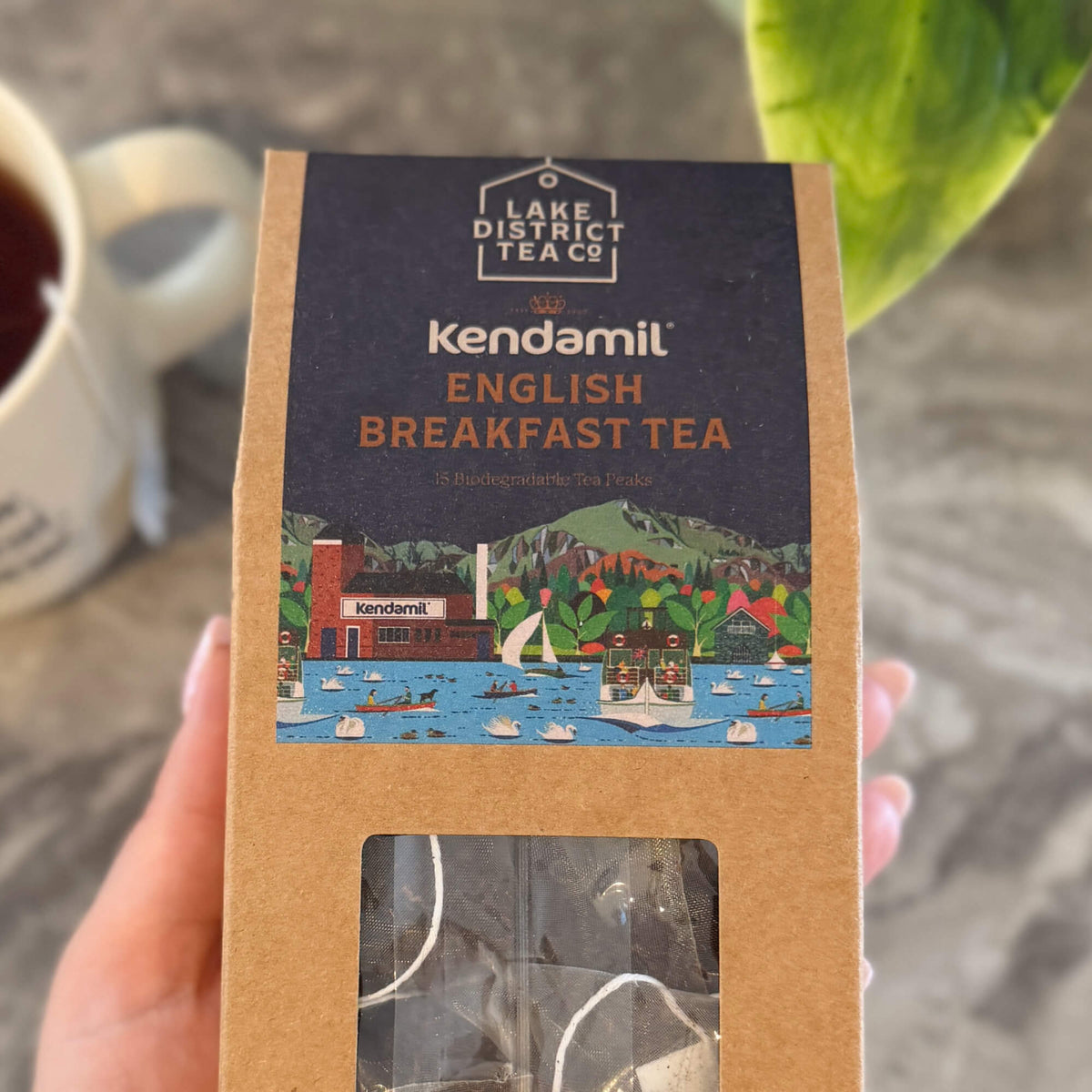 A hand holds a box of Kendamil x Lake District Breakfast Tea, which displays an illustrated lakeside scene and reveals tea bags inside. In the background, a cup of rich, full-bodied tea and green plant leaves complete the setting.