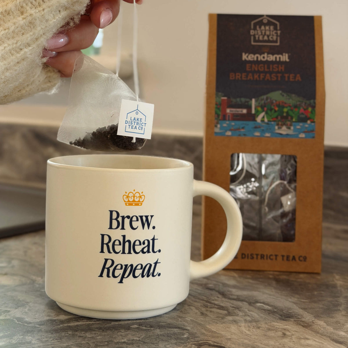 A hand dips a tea bag labeled Kendamil x Lake District Breakfast Tea into a mug that reads Brew. Reheat. Repeat. In the background, a box of Kendamil x Lake District Breakfast Tea—a full-bodied Assam, Ceylon, and Kenyan blend—sits on the kitchen counter.