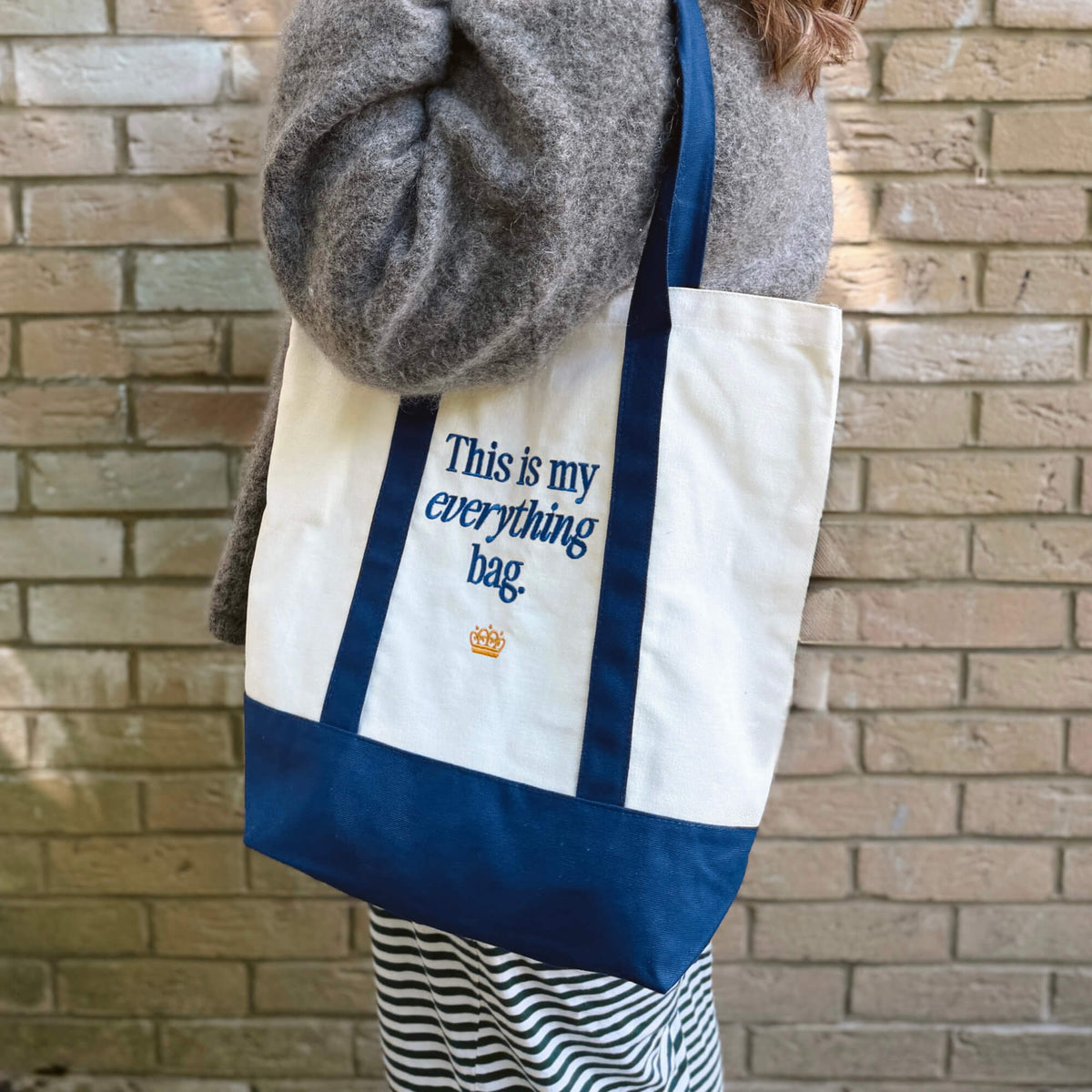 A person stands in front of a brick wall, wearing a gray sweater and striped skirt, holding the “This is my everything bag.” Tote Bag—a stylish white and blue tote featuring the phrase “This is my everything bag” and a small crown graphic. It’s perfect for family outings or carrying baby essentials.