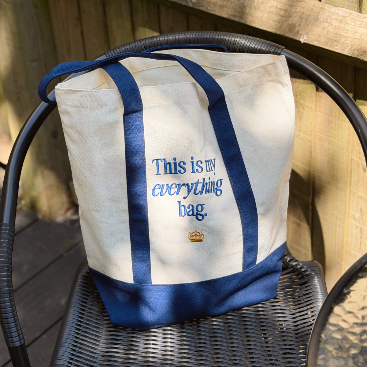 The “This is my everything bag.” Tote Bag, featuring a cream and blue design with blue handles and a small gold crown beneath the printed text, rests on a black chair. Ideal for carrying baby essentials or packing for family outings, sunlight casts soft shadows across the scene.