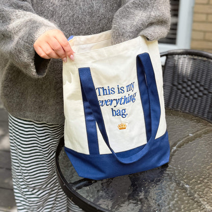 A person in a gray sweater and striped pants holds the “This is my everything bag.” Tote Bag, featuring cream and blue colors with the phrase “This is my everything bag” and a small crown graphic beneath the text, displayed on a glass table—perfect for family outings.